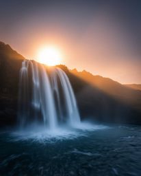A long-exposure photograph of a sun setting behind a waterfall, capturing motion and light trails. The image evokes a mystic,