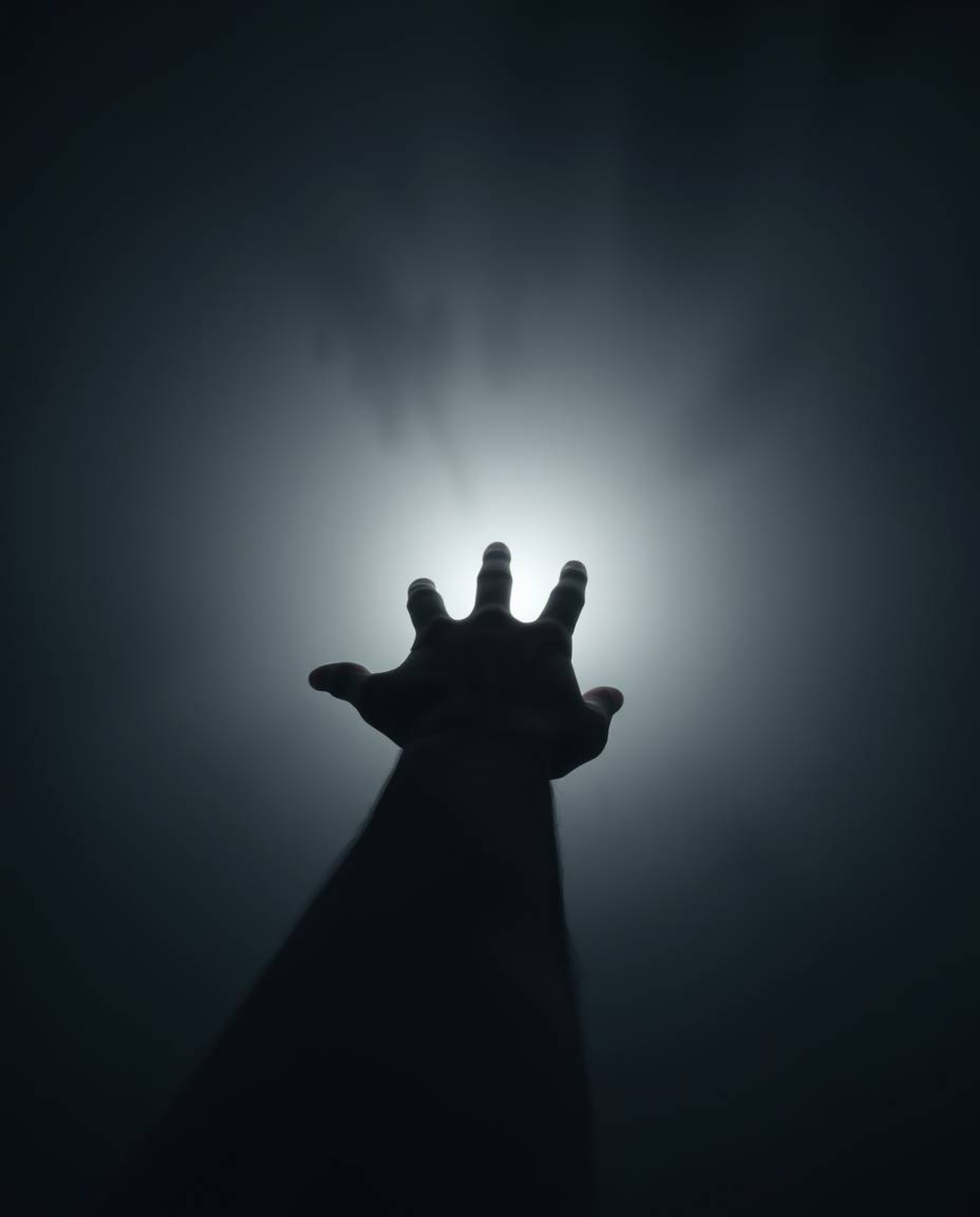This image captures a hand reaching towards a source of light from a darkened, fog-shrouded background. The moody style, low 