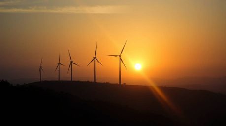 Capture the breathtaking beauty of a sunset behind a row of wind turbines on a hilltop in a cinematic style image.  Volumetri