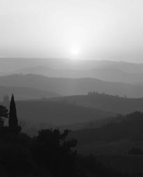 This is a classic black and white image depicting a timeless and elegant Tuscan sunset over misty, rolling hills. The scene i
