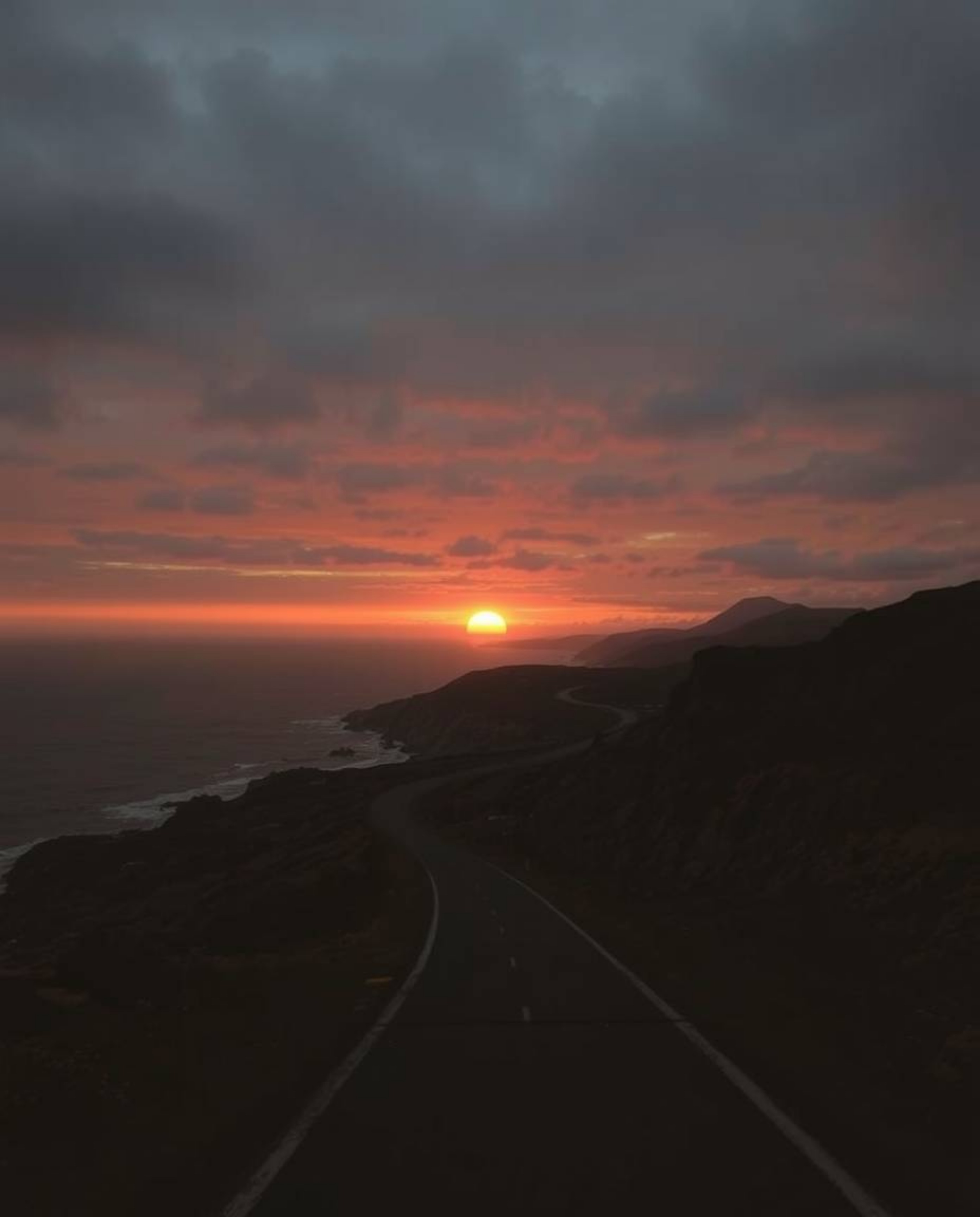 This image depicts a sunset sky over a winding coastal highway, characterized by an eerie and unsettling atmosphere. The scen