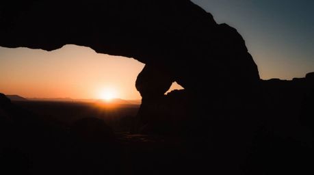 Capture the essence of a desert sunset framed by a natural rock arch with a moody and atmospheric photograph. This image emph