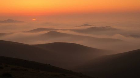 A captivating low-angle image showcases fog-covered hills bathed in the warm glow of the setting sun. Rendered in natural ear
