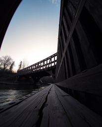 This is a low-angle architectural photograph capturing an old wooden bridge spanning a river at sunset. The wide-angle perspe