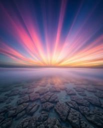 A long exposure photograph captures a colorful sky over a coral reef at low tide, featuring dramatic volumetric lighting. Vis