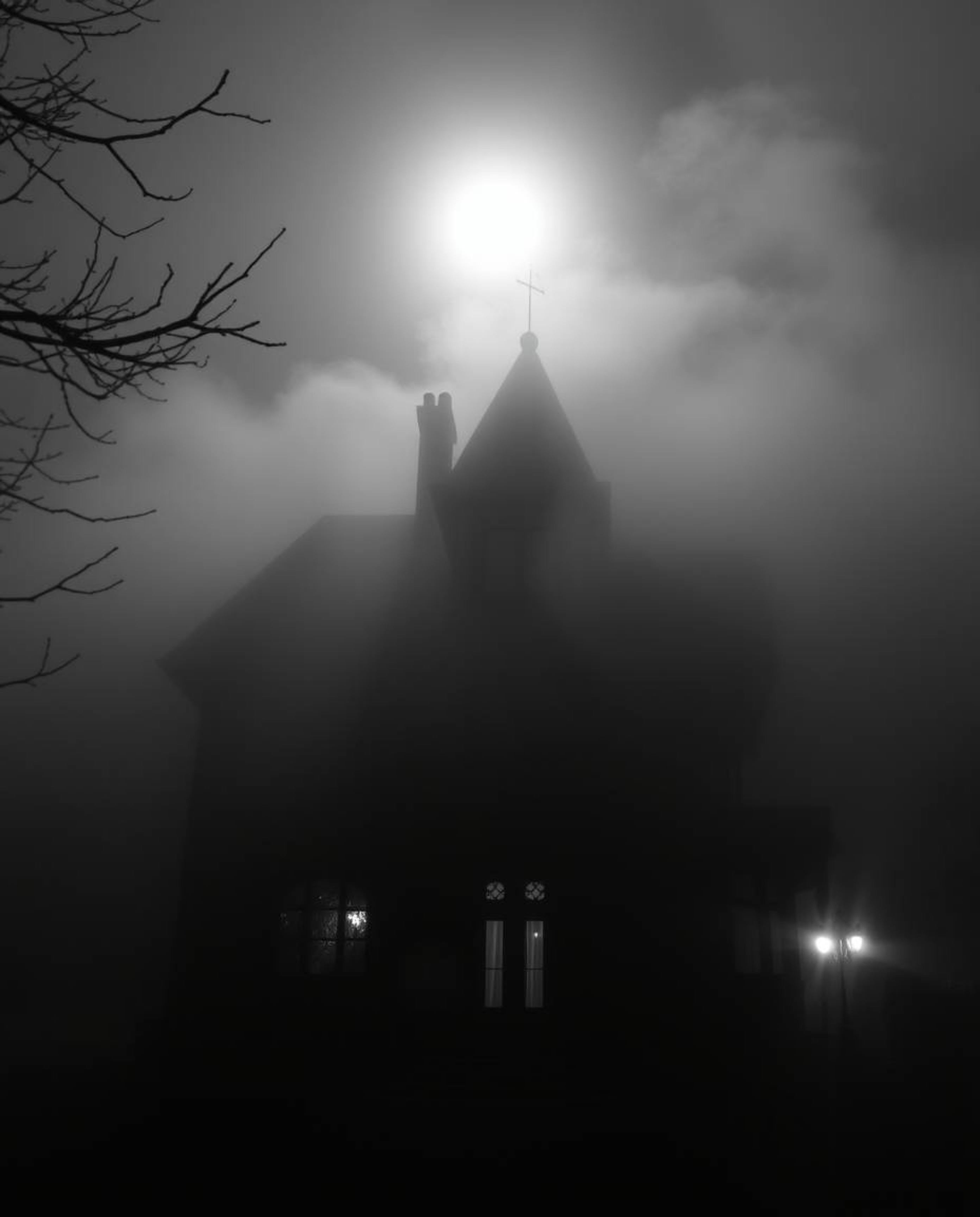 A classic black and white photograph capturing a fog-shrouded Victorian mansion bathed in moonlight. The image emphasizes tim