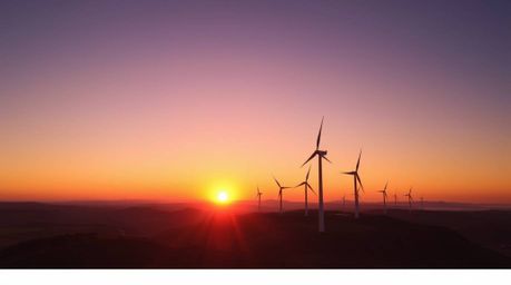 Capture the breathtaking beauty of a vibrant sunset painting the sky behind a row of wind turbines perched atop a scenic hill