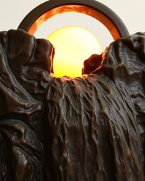 This is a cast bronze sculpture depicting a sun setting behind a dramatic waterfall. The artwork showcases a beautiful antiqu