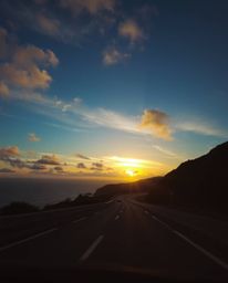 Captured from a low angle perspective, this photograph showcases a stunning sunset sky over a winding coastal highway. The sc