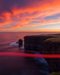 This is a long exposure photograph that captures a sunset over a coastal cliff, with the graceful motion of soaring birds. Th